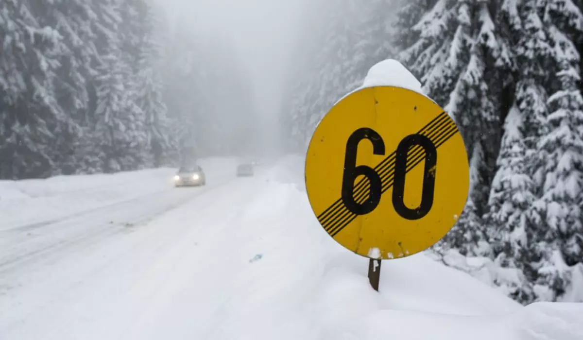Drum de munte acoperit cu zăpadă și un indicator rutier de culoare galbenă de încetare a limitării vitezei la 60 de kilometri pe oră