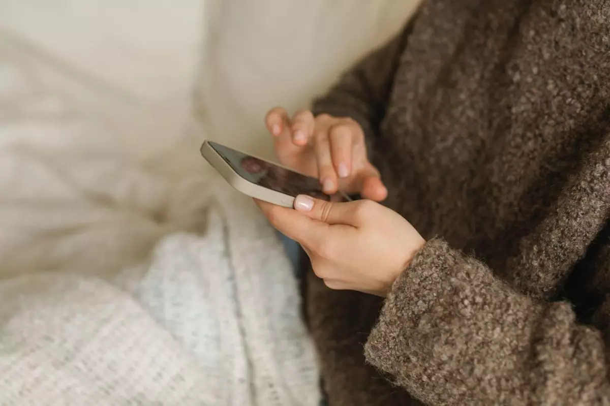 Prim plan al unei tinere cu un pulover tricotat care stă pe o canapea sub o pătură și își folosește telefonul mobil