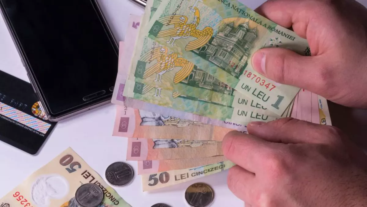 Un bărbat ține în mână mai mulți bani hârtii, iar pe masă mai sunt monede, un card bancar și un telefon.