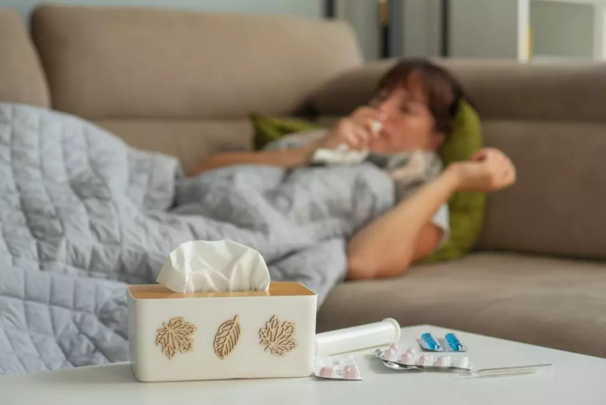 O femeie bolnavă de gripă este întinsă pe un pat se vede în fundal, iar în prim-plan este o masă cu medicamente și șervețele de nas. În România a fost declarată stare de alertă