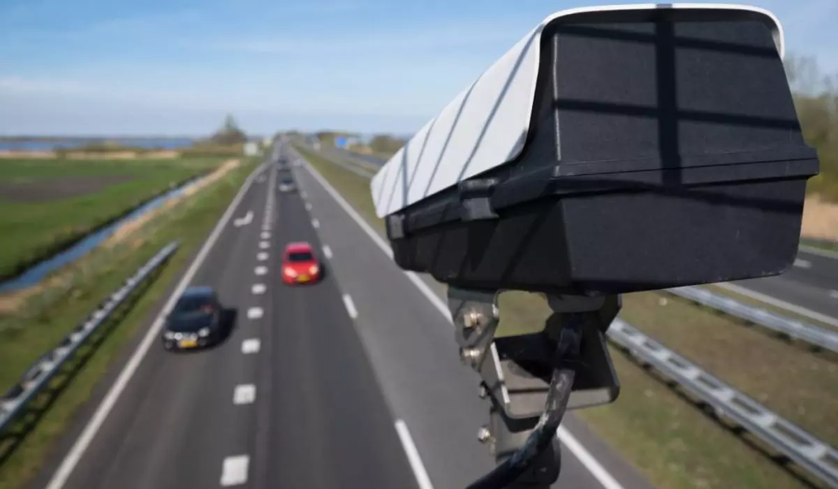 camera video care inregistreaza viteza din trafic. se observa o autostrada cu trei benzi pe sens pe care circula doua masini, una rosie si una neagra