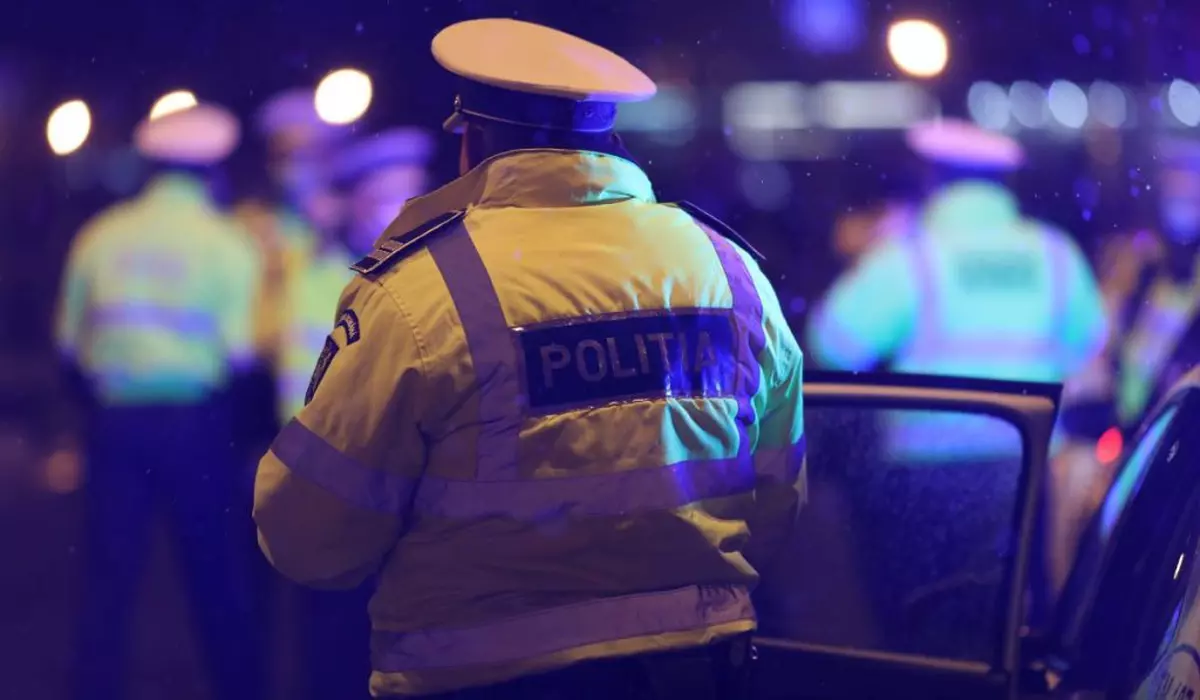 Polițist îmbrăcat în uniformă, fotografiat din spate, noaptea, lângă o mașină de Poliție cu ușa deschisă