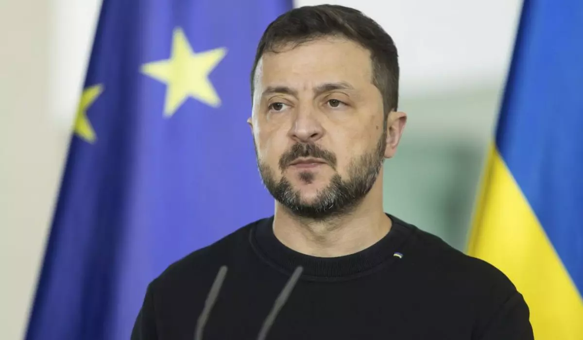 Volodimir Zelenski, președintele Ucrainei, într-o conferință de presă, îmbrăcat cu un pulover de culoare neagră, iar pe fundal steagurile Ucrainei și Uniunii Europene