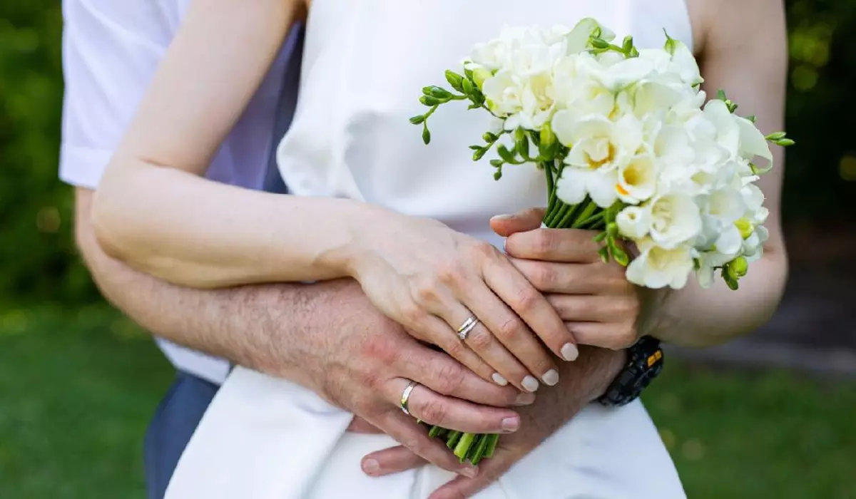 O mireasă și un mire se țin de mână și au un buchet de flori albe. Mireasa poartă o rochie albă, iar mirele poartă un costum. Scena este veselă și romantică, deoarece cuplul se îmbrățișează