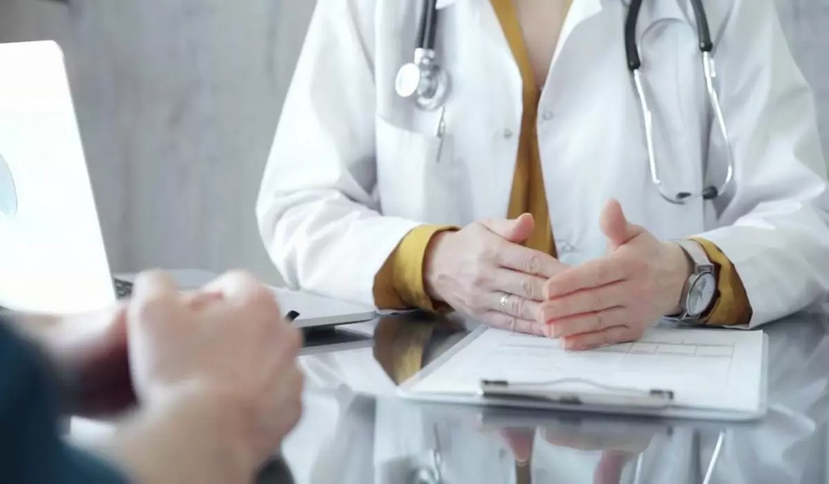 Medic îmbrăcat în halat alb, purtând un stetoscop în jurul gâtului și un ceas de mână, gesticulând ușor deasupra unei mese pe care se află o fișă medicală și un laptop deschis.