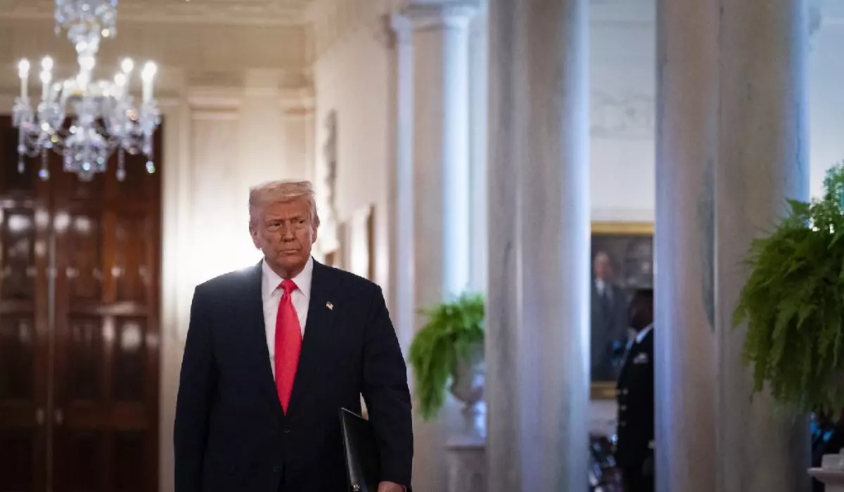 Donald Trump, îmbrăcat într-un costum albastru închis, cămașă albă și cravată roșie, mergând pe un hol. În dreapta lui sunt niste ferestre cu perdele albe, în stânga sus este un candelabru.