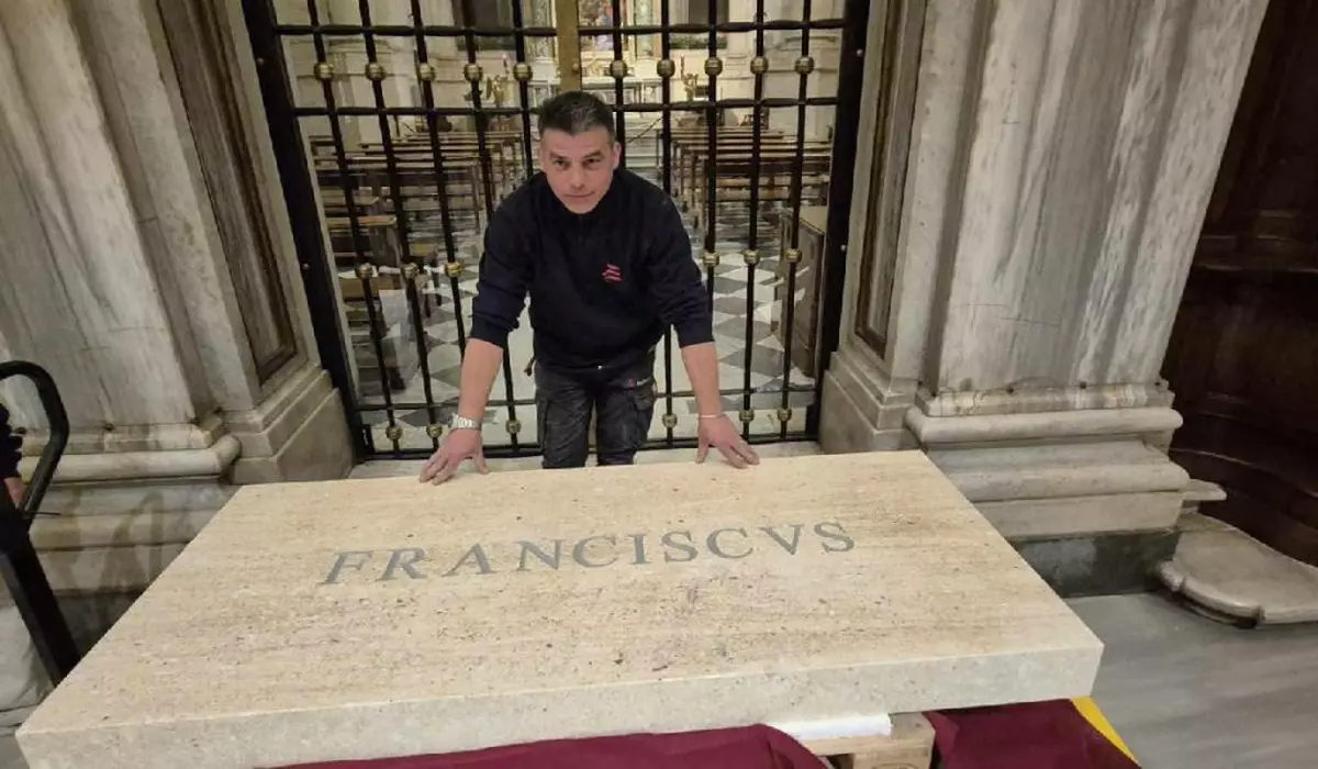 Un bărbat îmbrăcat în negru stă cu mâinile sprijinite pe o placă de marmură pe care este inscripționat „Franciscus”