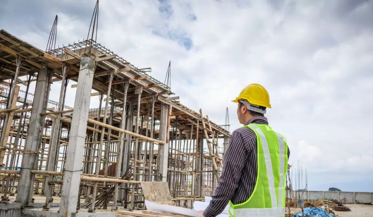 clădire impobiliară în construcții, privită de un inginer îmbrăcat în vestă reflectorizantă cu o cască de constructor pe cap