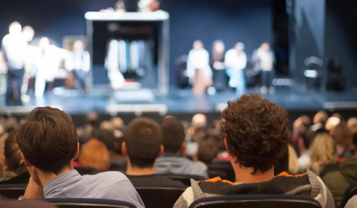 În imagine sunt ilustrați oameni care se uită la o piesă de teatru