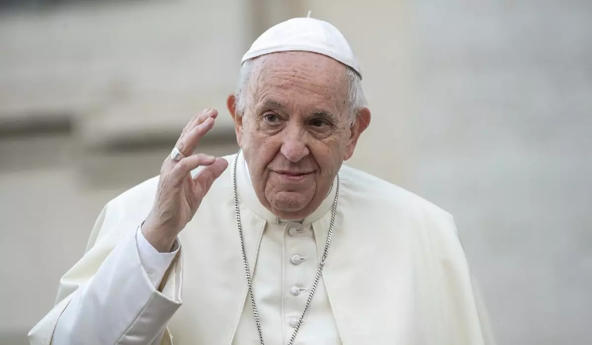 În imagine este ilustrat Papa Francisc, în timp ce ține mâna dreaptă ridicată