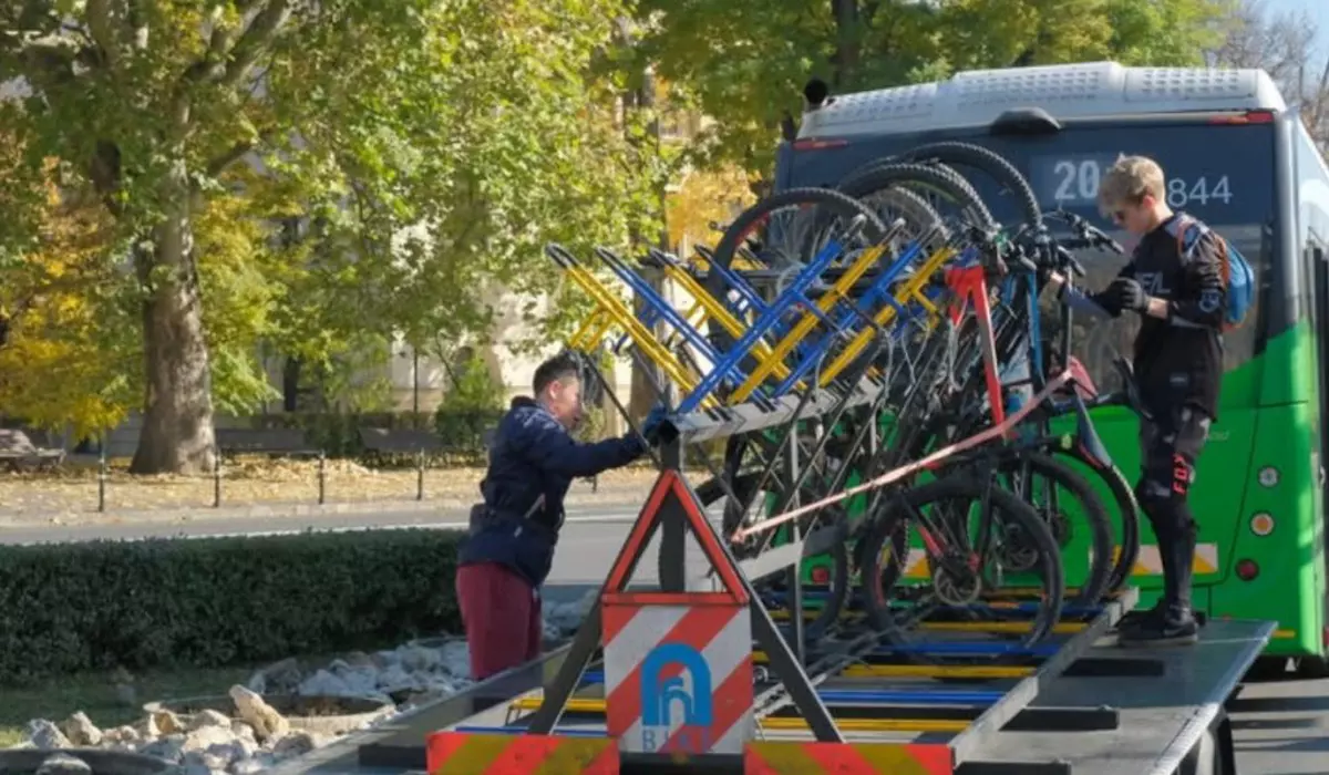 În imagine se observă un sistem de transport al bicicletelor montat în spatele unui autobuz, într-un peisaj urban. Două persoane, îmbrăcate în echipament sportiv, sunt ocupate cu încărcarea sau descărcarea bicicletelor de pe suportul special.
