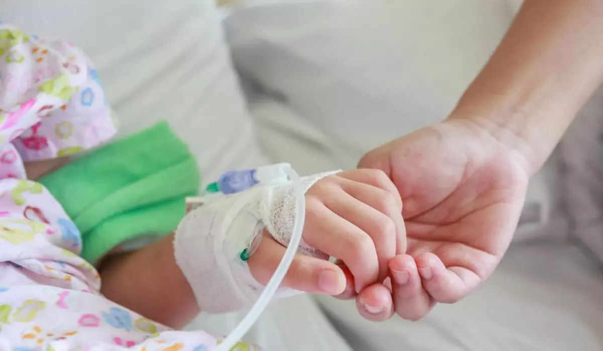 mama care ține de mână un copil în spital, cu o branulă în mână