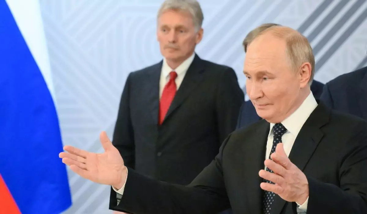 Putin cu brațele deschise, îmbrăcat în costum negru, camasa alba si cravata neagra. În spate, Dmitri Peskov, secretarul de presă al Kremlinului, în costum negru, cîmașă albă și cravată roșie. Președintele rus Vladimir Putin în timpul întâlnirii sale cu președintele egiptean Abdel Fattah El-Sisi în marja celui de-al 16-lea Summit BRICS de la Kazan.