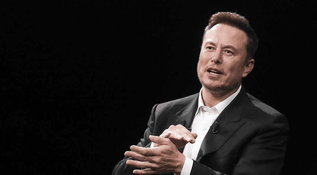 Imagine cu Elon Musk pe un Fundal negru, el îmbracat în costum gri închis, in timp ce vorbeste, făcând un semn cu mainile apropiate