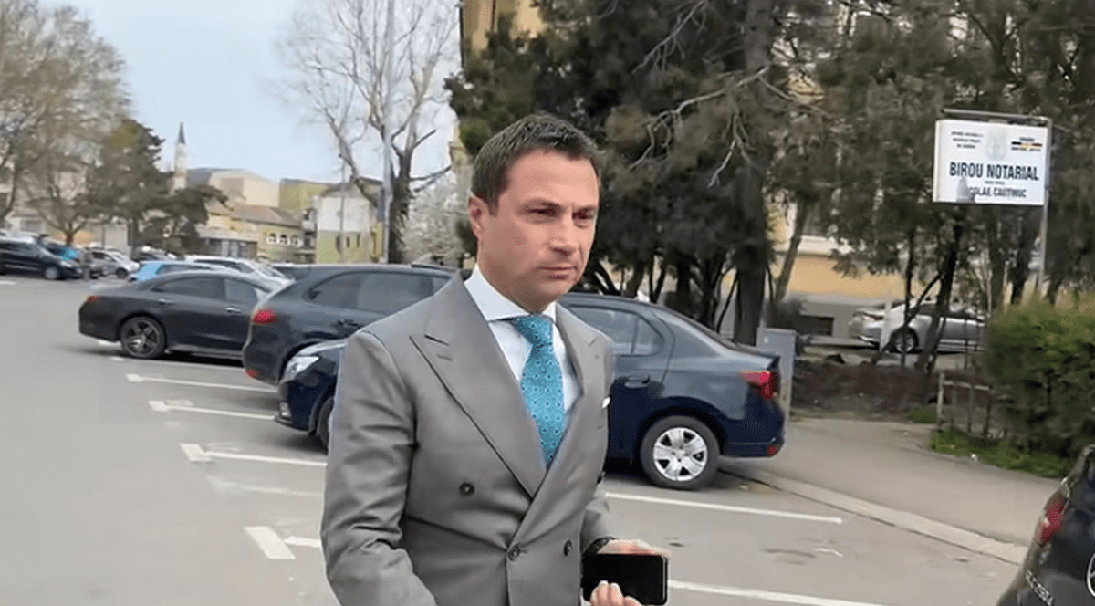 Radu Cristian merge pe stradă într-un costul gri și ținând în mâna dreaptă telefonul mobil, august 2025