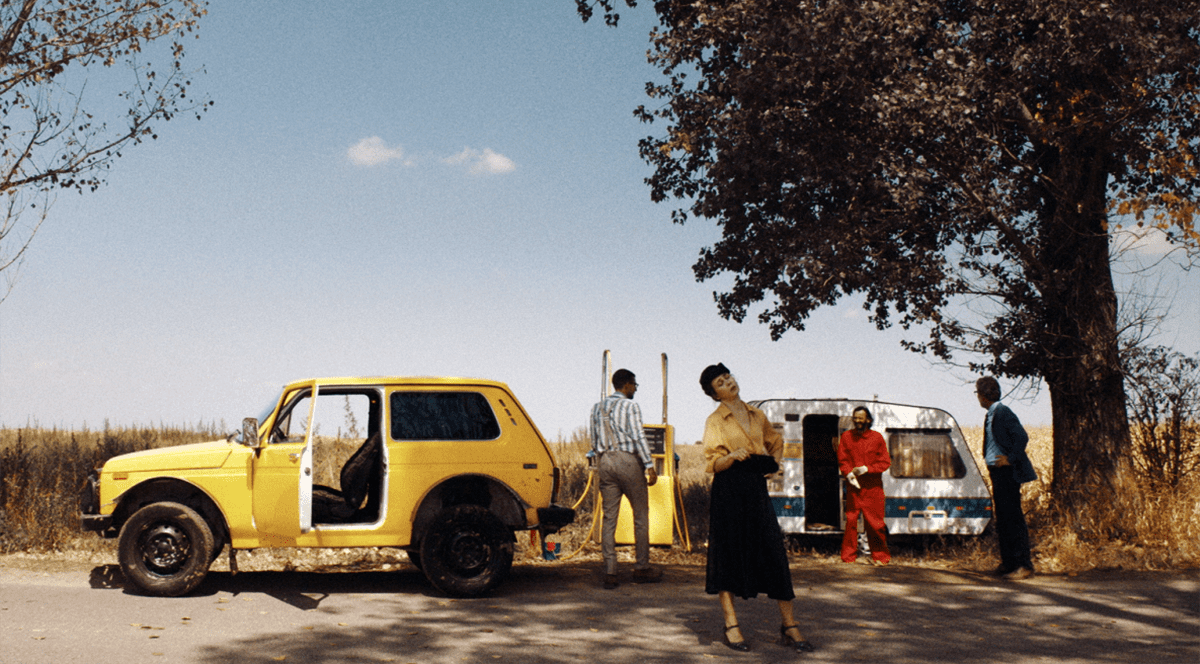 Imagine dintr-un film, o masina galbena si trei personaje, un barbat in alb, o femeie in galben si negru si una in rosu