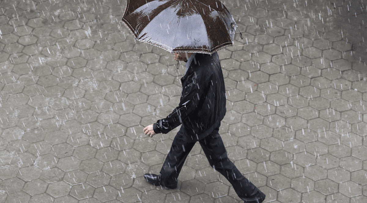 O persoană îmbrăcată în haine de culoare închisă merge prin ploaie pe un pavaj hexagonal, adăpostindu-se sub o umbrelă maro.