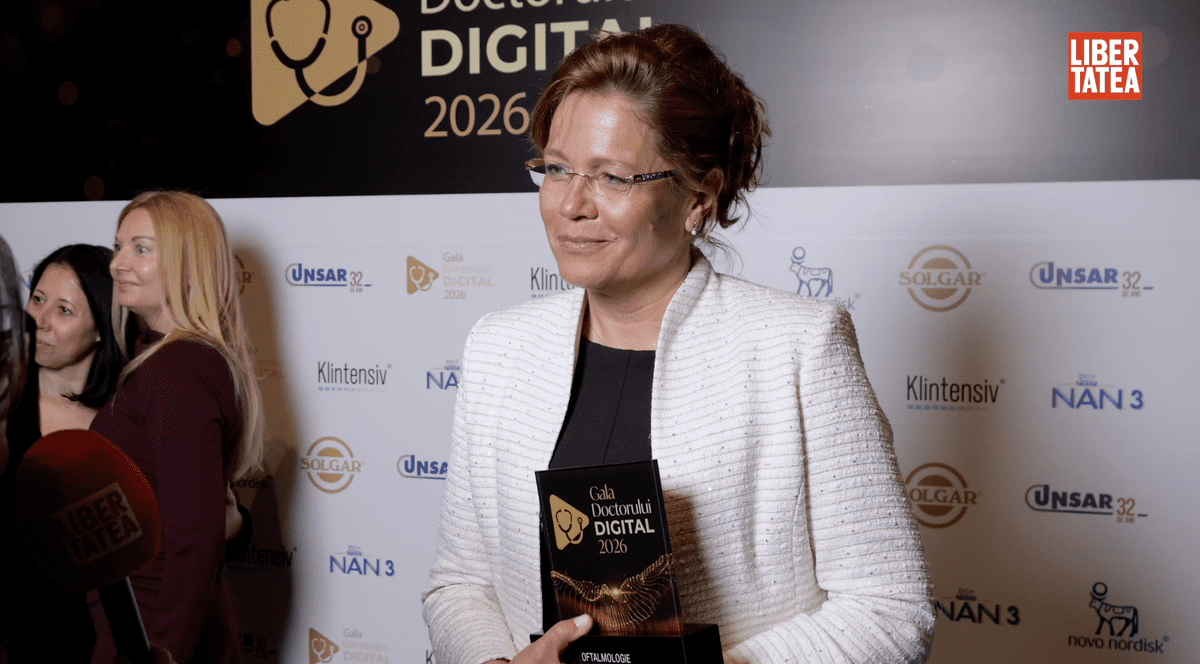 Dr. Andreea Ciubotaru, la Gala Doctorului Digital 2026, îmbrăcată cu o bluză neagră și un sacou alb, pe un fundal cu un panou alb și ținând în mână un premiu.