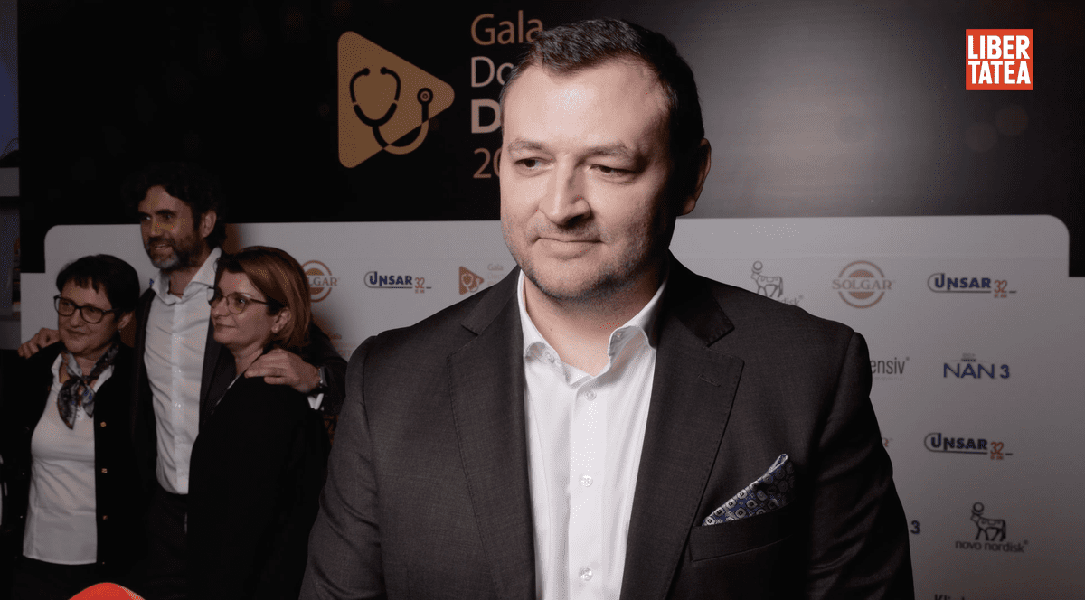 Dr. Ionuț Leahu, medic stomatolog, la Gala Doctorului Digital 2026, îmbrăcat cu o cămașă albă și un sacou nergu, pe un fundal cu un panou alb și alte trei persoane în stânga, spate, care stau asezate pentru a face o fotografie.