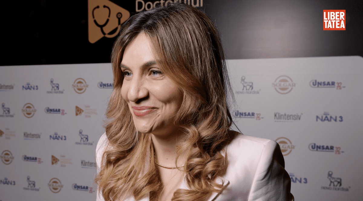 Dr. Monica Dima, medic specialist dermatolog, la Gala Doctorului Digital 2026, îmbrăcată cun un sacou roz pal și un lănțișor auriu la baza gâtului, pe un fundal cu un panou alb și negru.