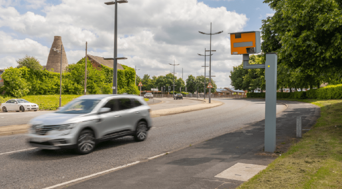 Imaginea prezintă un autoturism argintiu în mișcare pe o șosea, monitorizată de o cameră radar portocalie amplasată pe marginea drumului, într-o zonă cu vegetație și clădiri în fundal.