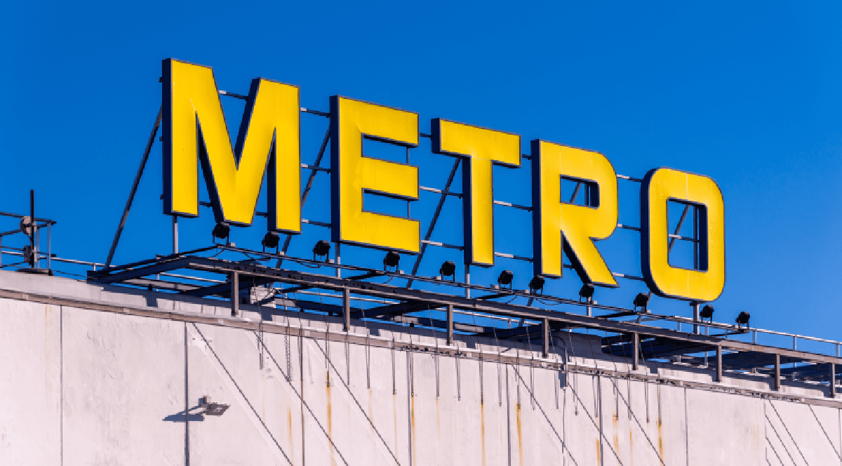 Imaginea prezintă sigla galbenă a magazinului Metro, amplasată pe acoperișul unei clădiri sub un cer de un albastru senin.