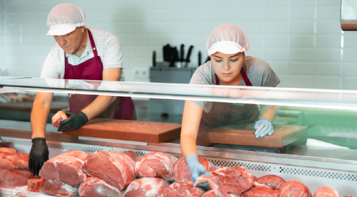 Imaginea surprinde doi măcelari purtând echipament de protecție sanitară în timp ce aranjează și pregătesc diverse sortimente de carne proaspătă în vitrina frigorifică a unui magazin de specialitate.