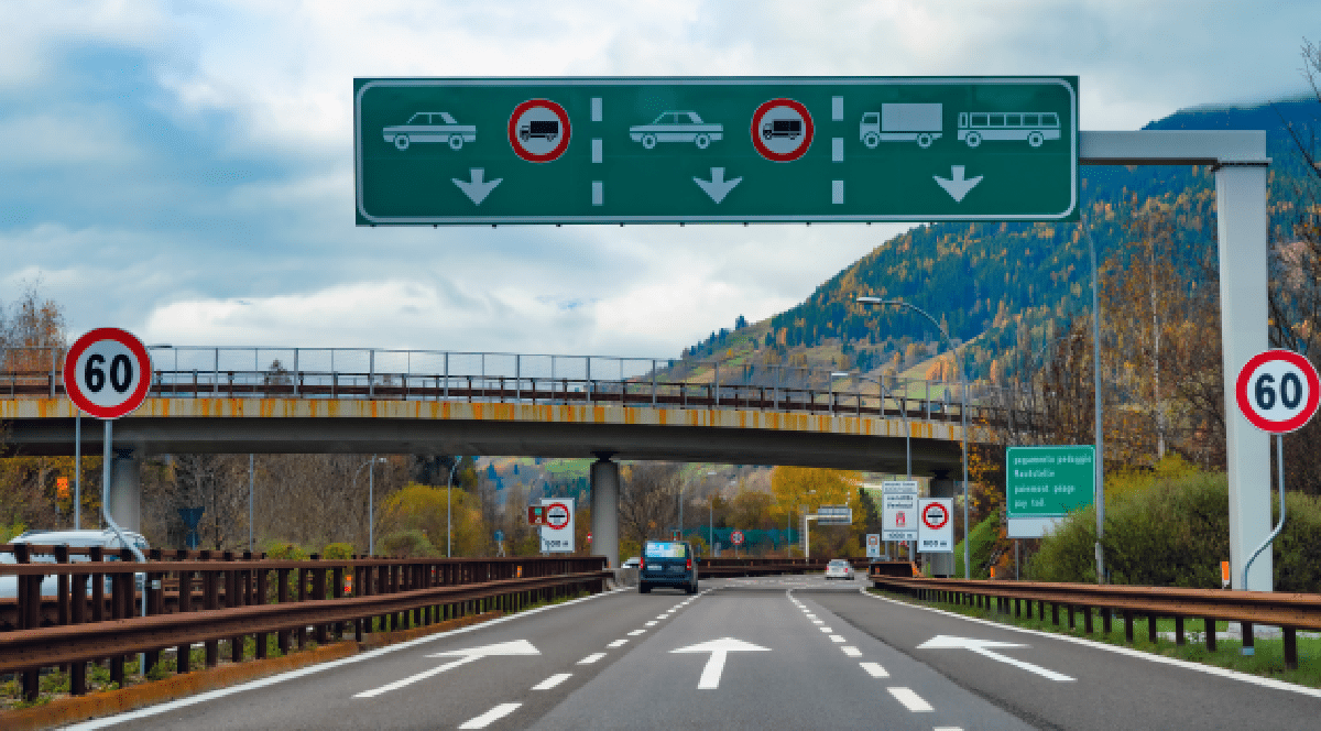 Imaginea prezintă o secțiune de autostradă cu indicatoare rutiere și panouri verzi care direcționează traficul pe benzi în funcție de tipul vehiculului, având pe fundal un peisaj montan.