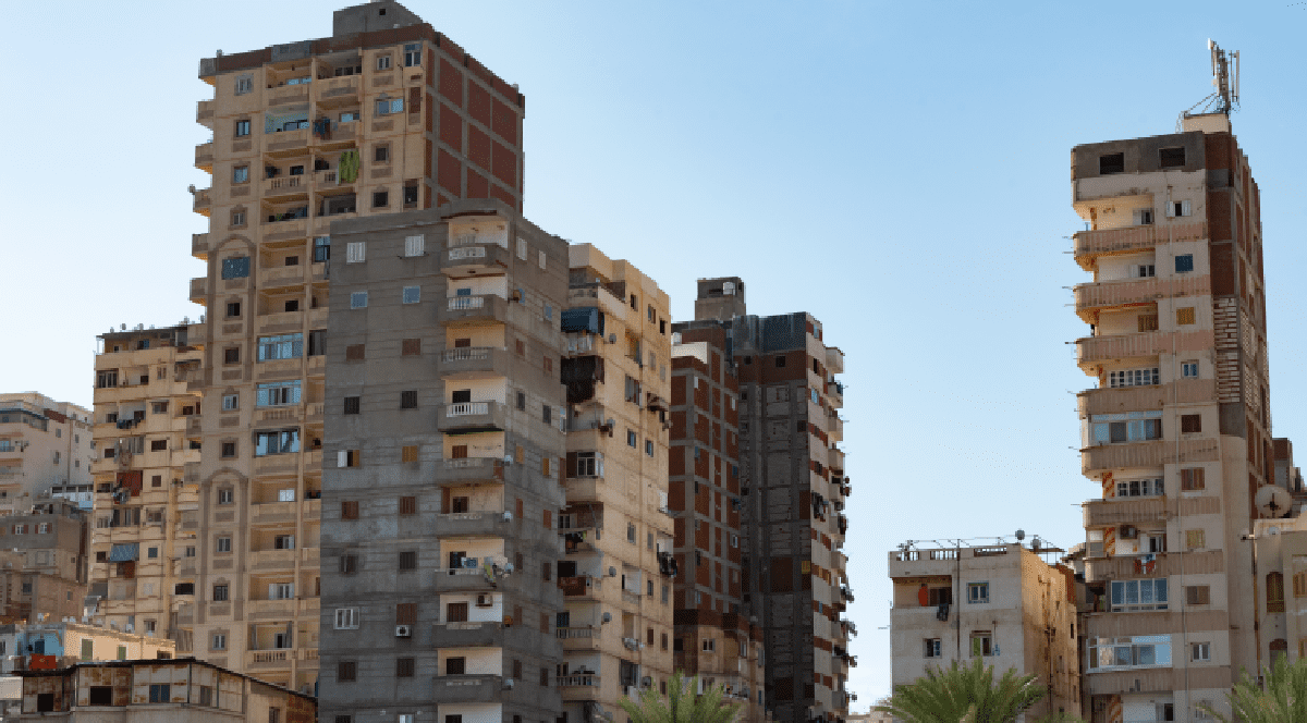 Imaginea prezintă un peisaj urban dens, cu blocuri de apartamente înalte construite în stiluri variate, printre care se zăresc câțiva palmieri sub un cer senin.