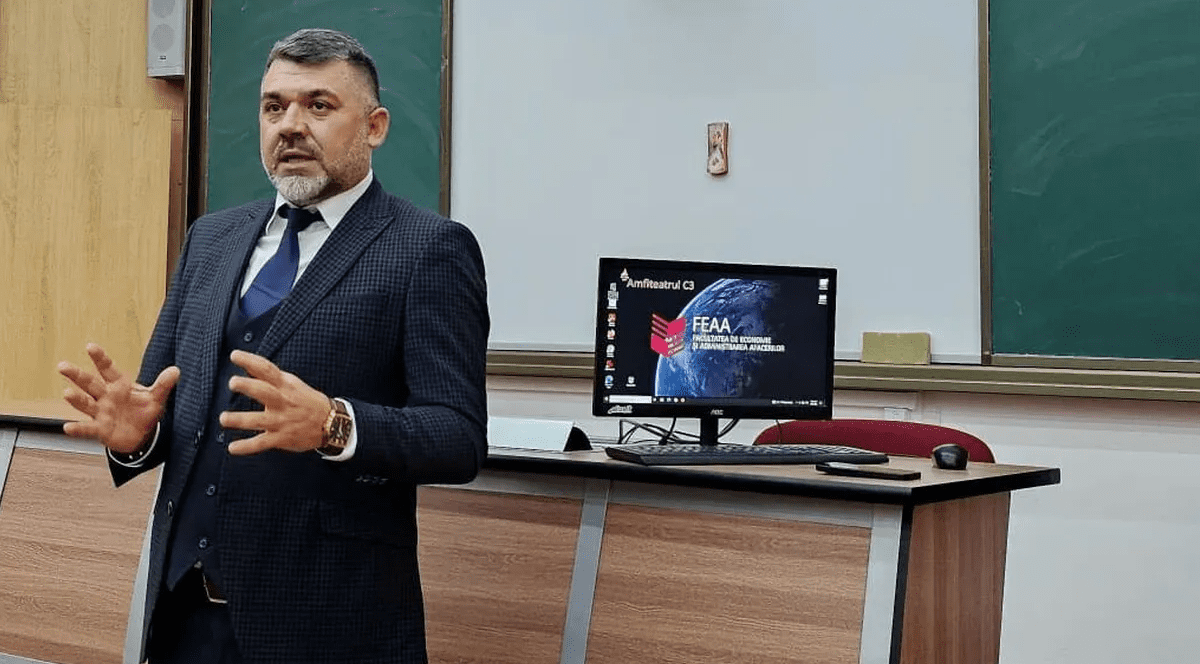 Costel Grojdea predă studenșilor fiind îmbrăcat într-un costum de culoare închisă, asortat cu o cravată iar în spatele său se află un ecran. Foto: Facebook- Costel Grojdea