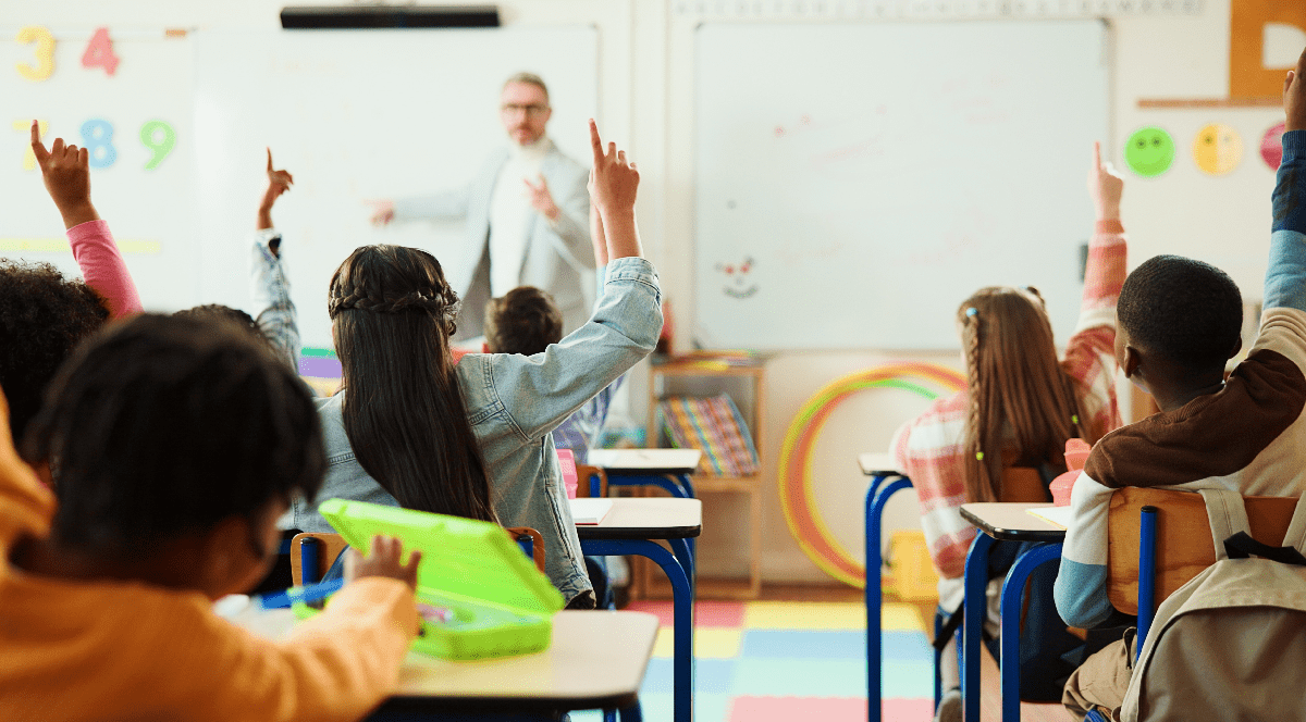 Imaginea prezintă o sală de clasă plină de elevi care ridică mâna pentru a răspunde, în timp ce un profesor le vorbește din fața tablei.