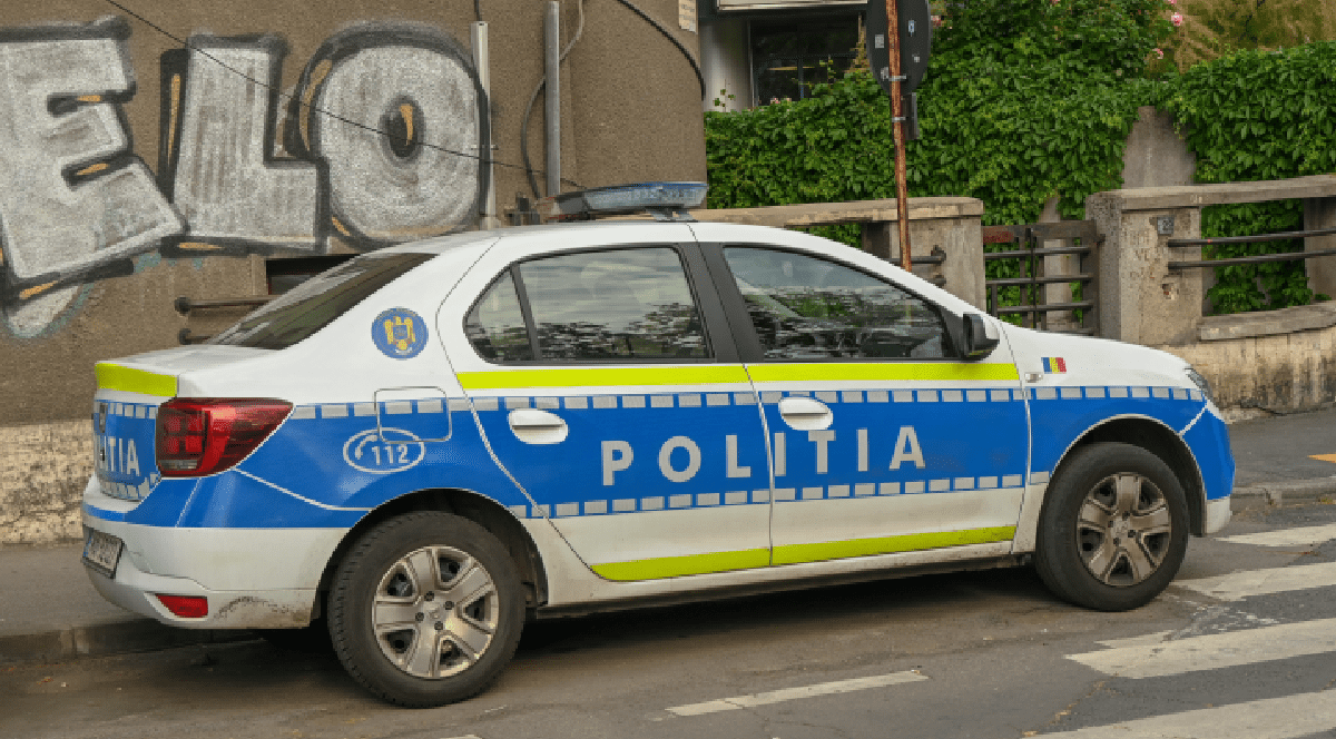 Imaginea prezintă un autoturism de poliție din România parcat pe marginea unei străzi, lângă o trecere de pietoni și un perete cu grafiti.