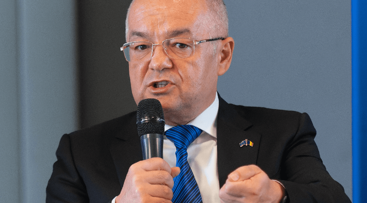 Imaginea prezintă un prim-plan al politicianului Emil Boc, purtând ochelari și costum, în timp ce vorbește la un microfon și gesticulează.