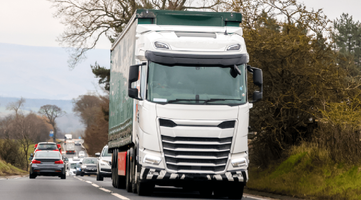 O imagine dinamică ce surprinde un camion alb cu remorcă verde circulând pe o șosea cu două benzi, fiind urmat de o coloană de autoturisme într-un peisaj rural.