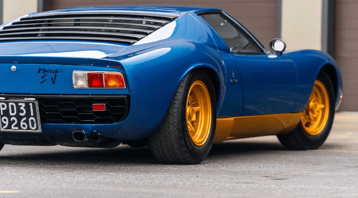 Imaginea prezintă partea din spate a unui automobil clasic Lamborghini Miura SV de culoare albastră, echipat cu jante aurii, parcat în fața unei clădiri cu uși de garaj.