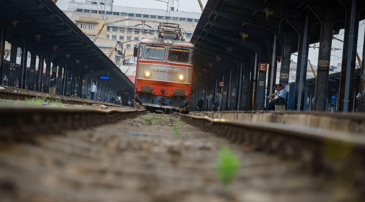 Imaginea surprinde o perspectivă de la nivelul șinelor asupra unei locomotive românești care intră în gară, sub acoperișul peroanelor flancate de coloane metalice.