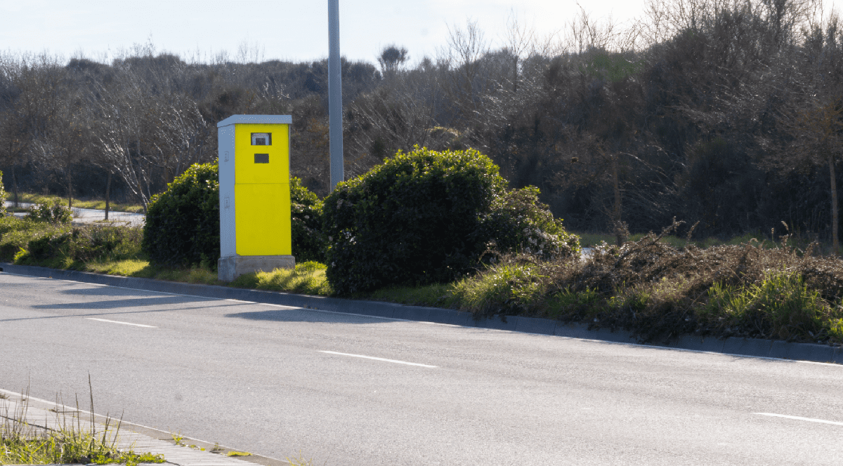 Imaginea prezintă o cabină galbenă de radar amplasată pe marginea unei șosele, fiind flancată de tufișuri verzi.