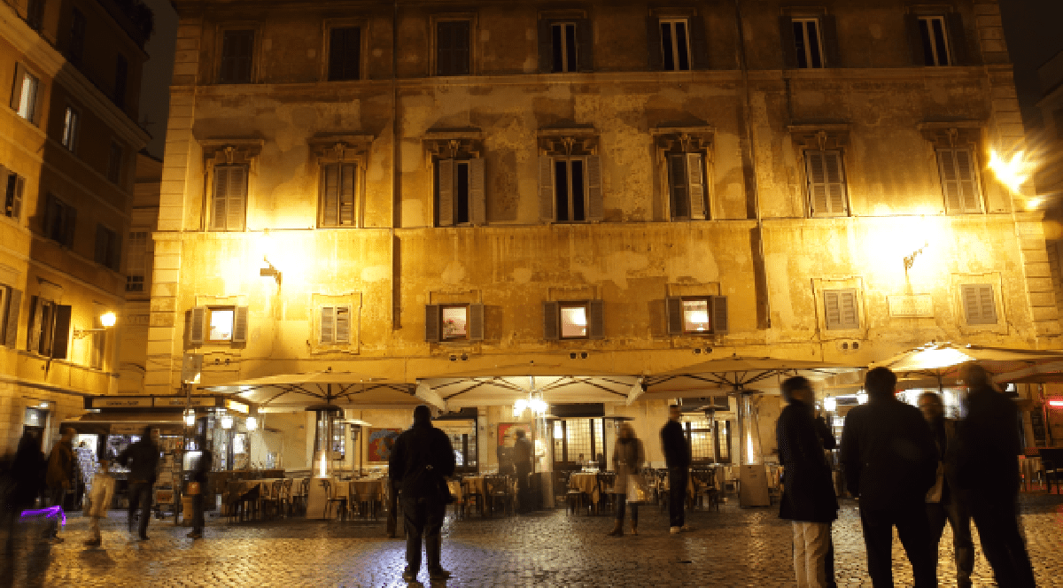 Imaginea surprinde atmosfera nocturnă a unei piețe din Roma, unde trecătorii și clienții se plimbă pe lângă terasele iluminate ale restaurantelor, aflate la parterul unei clădiri istorice cu fațadă veche.