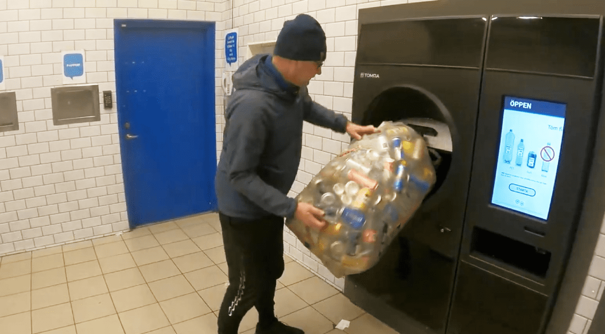 Imaginea surprinde un bărbat care golește un sac mare plin cu doze de aluminiu într-un automat industrial de colectare și reciclare a ambalajelor, într-un spațiu special amenajat.