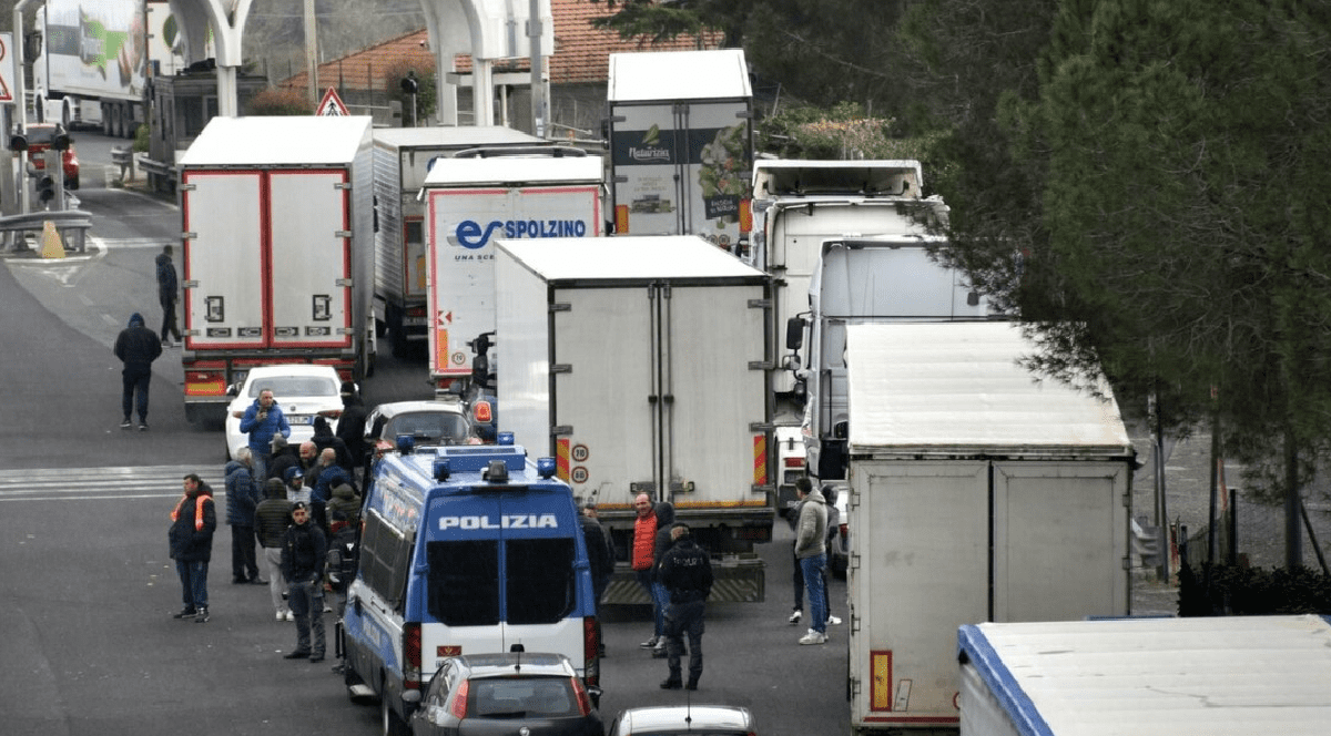 Imaginea surprinde o coloană de camioane blocate la un punct de taxare rutieră, unde mai mulți ofițeri de poliție și o autoutilitară „Polizia” sunt prezenți în mijlocul unui grup de persoane, sugerând o acțiune de control sau un protest.