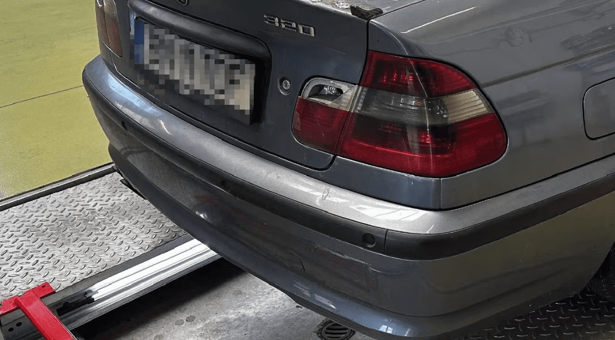 Imaginea prezintă partea din spate a unui autoturism BMW model 320 de culoare gri, aflat într-un atelier auto.