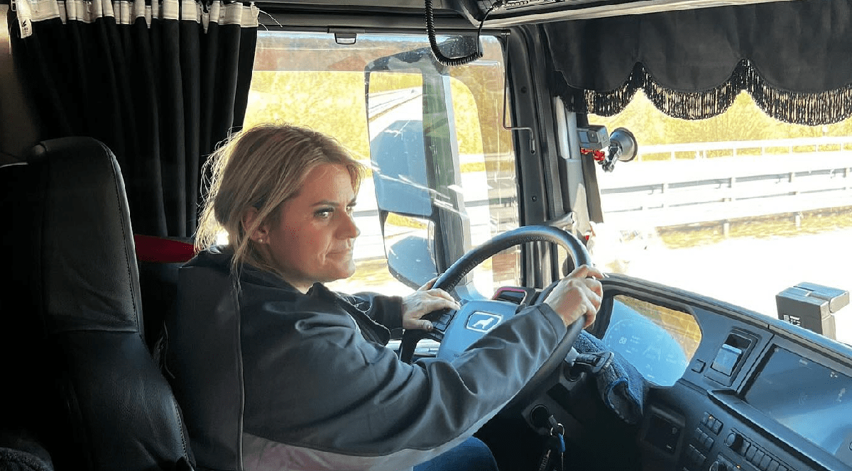 Imaginea prezintă o femeie aflată la volanul unui camion, surprinsă din interiorul cabinei în timp ce conduce pe un drum, într-o zi însorită.