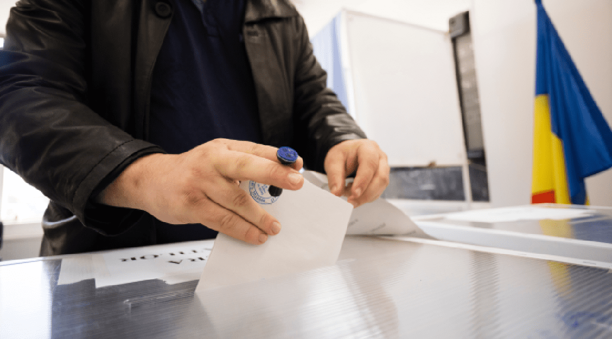 Imaginea surprinde un cetățean care introduce un buletin de vot ștampilat într-o urnă, în timpul unui proces electoral desfășurat în România, având steagul național vizibil în fundal.