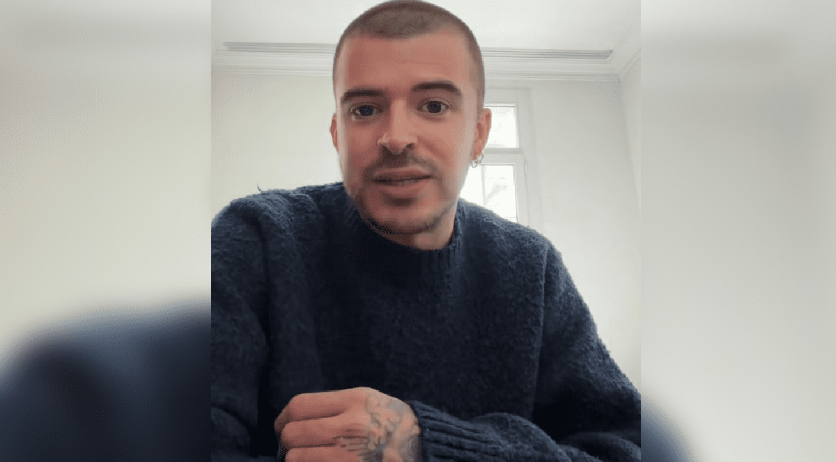 Imaginea surprinde un tânăr cu părul tuns foarte scurt și mâna tatuată, purtând un pulover albastru închis, în timp ce pare să vorbească în fața unei camere într-o încăpere luminoasă.