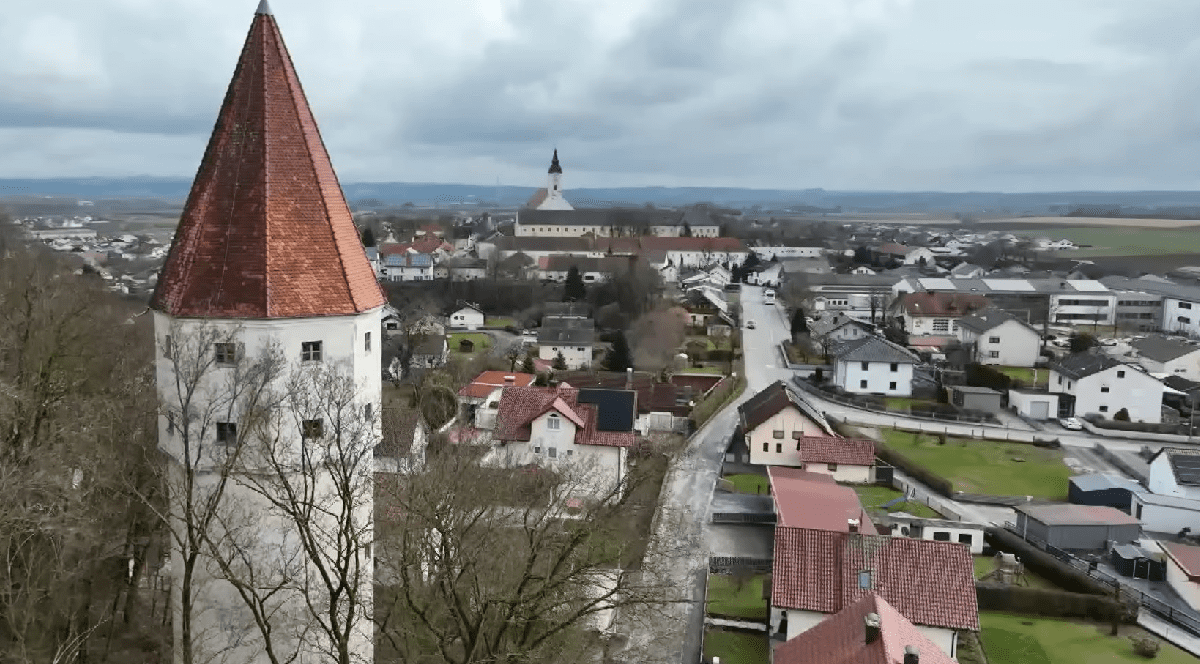 Imaginea oferă o vedere panoramică asupra unui orășel pitoresc, evidențiind un turn alb cu acoperiș conic din țiglă roșie în prim-plan.