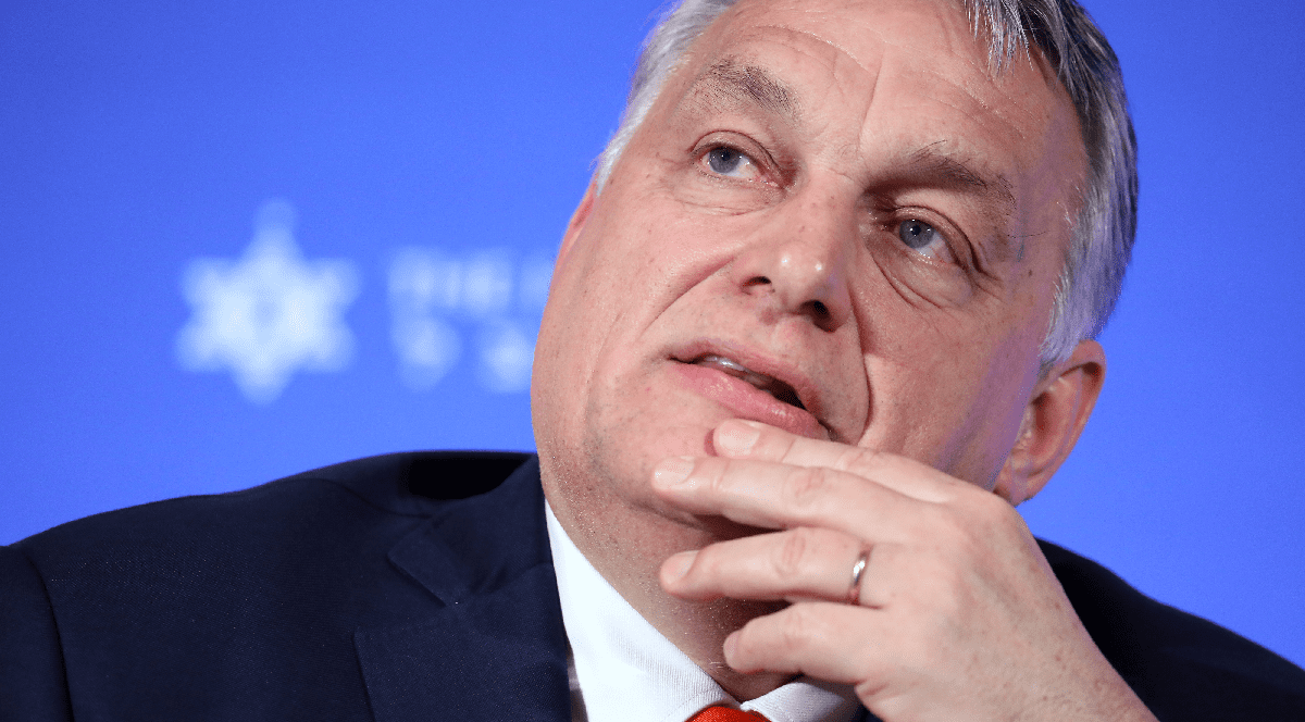 Imaginea prezintă un portret în prim-plan al politicianului Viktor Orbán, îmbrăcat în costum, cu mâna la bărbie, pe un fundal albastru.