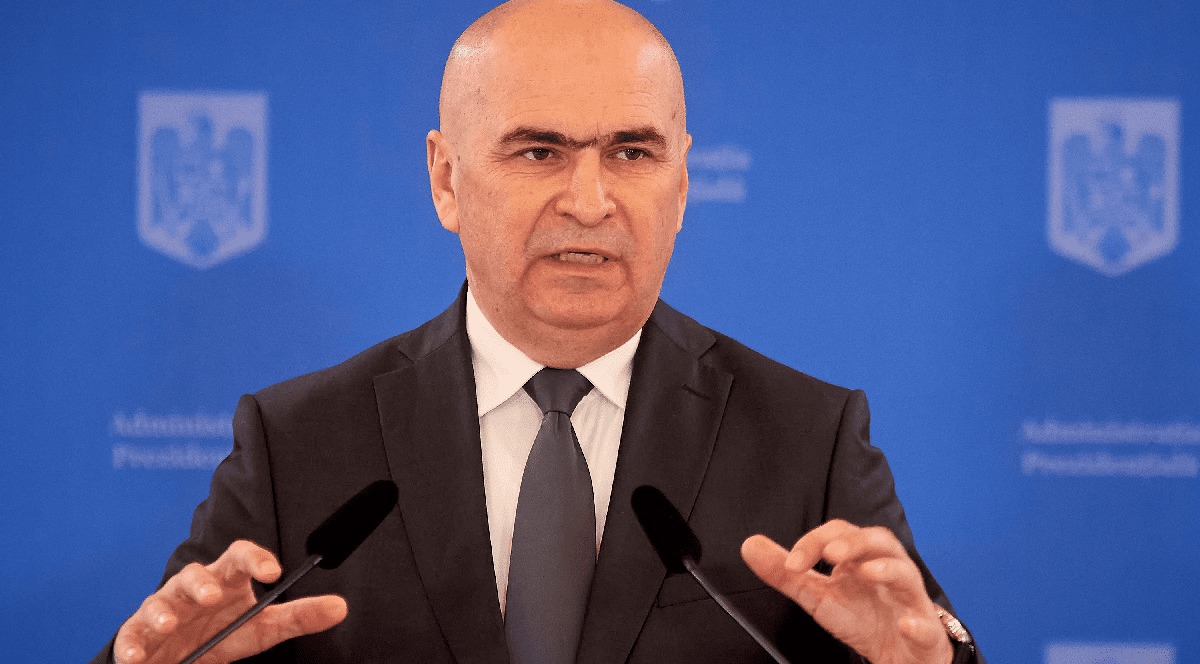 Imaginea îl înfățișează pe Ilie Bolojan susținând o declarație de la o tribună, gesticulând cu ambele mâini în fața unui fundal albastru ce poartă însemnele Administrației Prezidențiale.