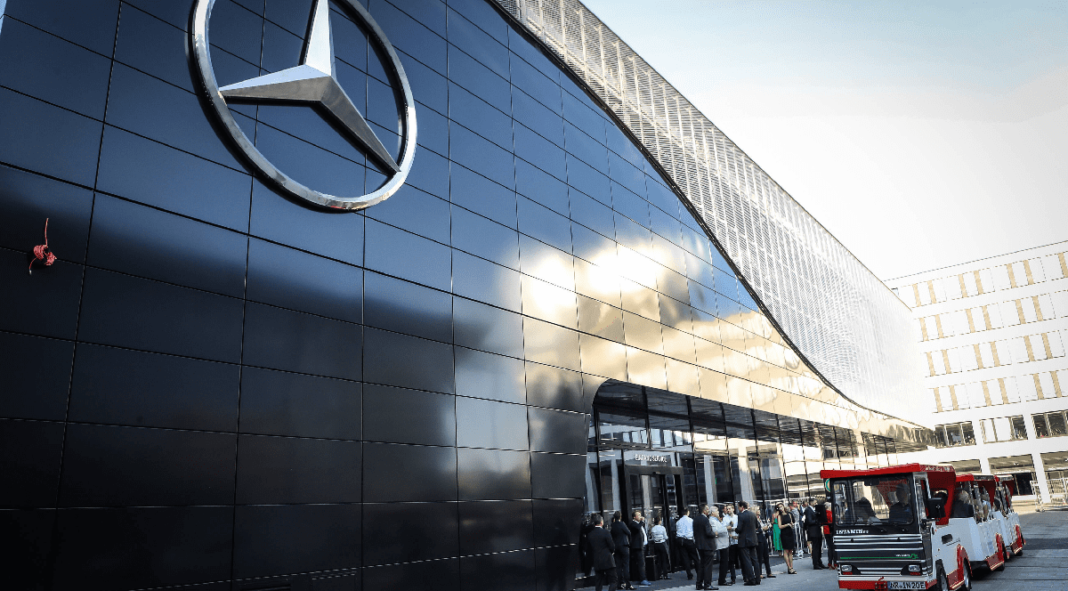 Imaginea prezintă fațada unei clădiri Mercedes-Benz, placată cu panouri negre lucioase și având logoul mărcii la dimensiuni mari, în fața căreia se află un grup de persoane.