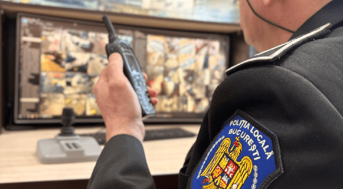 Imaginea înfățișează, din profil, un ofițer al Poliției Locale București care monitorizează mai multe ecrane de supraveghere video în timp ce utilizează o stație de emisie-recepție.