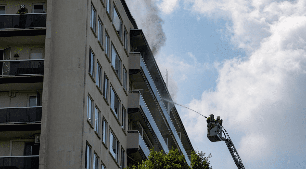 intervenția pompierilor la un incendiu izbucnit la un bloc de apartamente cu mai multe etaje. Din zona balcoanelor aflate la nivelurile superioare se ridică o coloană groasă de fum. În partea dreaptă a cadrului, doi pompieri aflați în nacela unei scări mobile de intervenție direcționează un jet puternic de apă spre balcoanele afectate. Pe partea stângă a clădirii, pe un balcon de la un etaj superior, stă un alt pompier echipat cu cască de protecție. În partea de jos a imaginii se pot distinge vârfurile unor copaci.